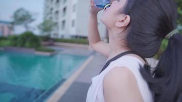 Aziatische jonge sportieve vrouw die een pauze neemt tijdens de training drinkwater, verfrissende bronnen, openbare recreatiefaciliteit zwembad ontspannen na het sporten, gezonde levensstijl vitamine mineraalwater, zijaanzicht video