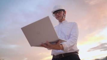 un ingénieur asiatique porte un casque de protection de sécurité travaillant avec un ordinateur portable sur un chantier de construction vide, une licence d'ingénierie, une analyse de la planification de la structure, un travailleur acharné travaille au fil du temps pendant le coucher du soleil