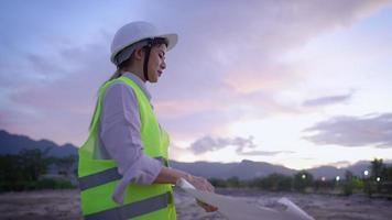 Young tired female construction site worker folding a blueprint paper while standing in safety workwear on muddy empty plot in evening time, over time working, dedicated employee or workaholic concept video