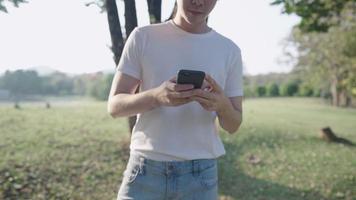 un hombre asiático con camisa blanca de ropa casual que usa un teléfono inteligente mientras camina dentro de un parque verde, las manos usan tecnología inalámbrica, actividad de ocio relajante, internet inalámbrico, esperando bajo la sombra de un árbol video