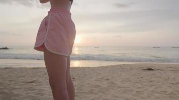 jovem garota asiática forte de cócoras na praia fazendo rotina de exercícios ao ar livre, agachamento de peso corporal na costa da praia de areia e pôr do sol no fundo, exercício matinal na praia, motivação para o exercício video