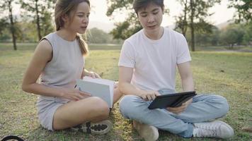 Young college friends sit down on the ground inside the park, discussing homework group project using digital tablet, friend brainstorm working together, relaxing meeting environment warm sunlight video