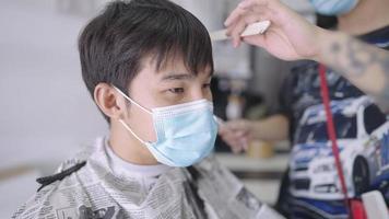 Attractive asian man wearing protective mask and friendly talking to male hipster hairdresser, reopening business, professional hairdresser designing a hairstyle and cutting with scissor and comb, video