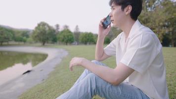 Young attractive asian man talking on smartphone while enjoying natural green park view during a day, wireless mobile technology devices, a usage of long-distance media, human society and environment
