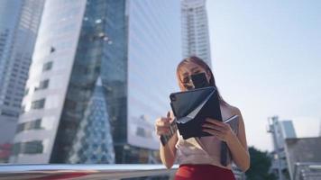 Asian attractive businesswoman wearing mask walking using digital tablet outside of a modern office building, self care during covid 19 coronavirus pandemic, office worker and convenience technology video