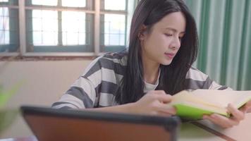 Young asia girl reading book at home table with tablet on the side, focusing concentrating on study, college student reviewing for exam test, self discipline general knowledge, free time leisure