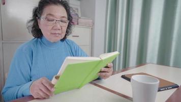 grand-mère âgée active asiatique en chandail froid bleu lit seul livre à la maison table de salon confortable, apprentissage de la vieillesse, sagesse et connaissances des gens, étude personnelle, vie de retraite heureuse, apprentissage continu