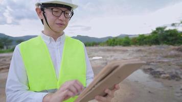 asiatisk ingenjör arbetare bär glasögon och skyddande arbetskläder, står på utomhusbyggarbetsplatsen, specialistarbete, tittar på arkitekturritning, analysidé och planeringsframsteg video
