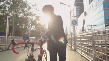 Aziatische zelfverzekerde werknemer die zijn fiets duwt op het viaduct van de stad, het bedrijfsleven en het milieu van transportconcept, zonreflectie op glas modern gebouw op de achtergrond, stedelijk landschap video