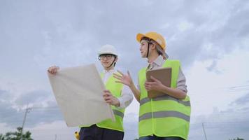 Asian workers wearing safety working gear standing outside construction field, surveying and design on building structure project, unfold blueprint drawing paper, teamwork coordinator planner video