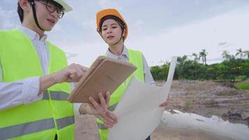 trabalhadores da construção civil de engenheiros asiáticos discutindo sobre papel de projeto de planta de construção civil, procurando verificar com pequenos detalhes com tablet, brainstorm de trabalho em equipe cooperando, luz natural do dia video