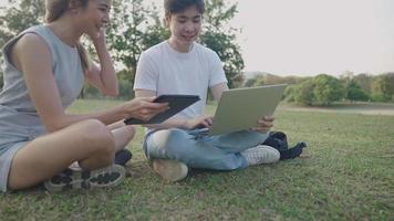 Two young genders diversity people sitting on a green meadow inside public park, happy teamwork having a conversation, remote working concept, ideas inspired by natural workplace, smart technology use video