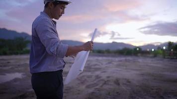 Aziatische jonge mannelijke ingenieur die een blauwdruk rolt, maak je klaar om naar huis te gaan, met uitzicht op de bewolkte avondhemel bovenop de grote bergen, toegewijde werknemer die laat in de avond, na verloop van tijd of concept werkt, video