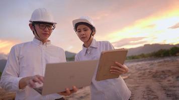 ein professioneller architekten-konstruktor-mentor, der einer jungen praktikantin auf einem laptop-computer-tablet am arbeitsplatz im freien, immobilienentwicklung, mit schutzhelm, bei sonnenuntergang eine joberklärung gibt video