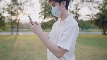 Young man wears protective face mask using cellphone walking alone inside the park at sunset, new normal distancing concept, wearing protective facemask when going outside in the new normal, relaxing