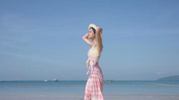 jovem e bela mulher asiática em roupas de verão com chapéu andando na praia com um mar no fundo. menina bonita de férias na ilha tropical. vento sopra ar fresco, chapéu de vento em câmera lenta