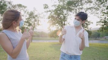 Young asian friends thumbs up showing confident, protecting themselves from transmitted infectious diseases virus, young couple wearing medical face mask standing inside the park new normal distancing video