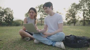 Young adult friends enjoying working together on the outdoor field trip, sit down on the ground green grass inside the park, discussing homework project, friends using laptop brainstorming on project video