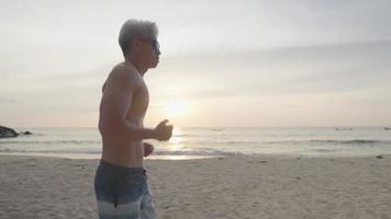 fuerte musculoso piel bronceada hombre de pelo gris usa gafas de sol corre en la playa con la luz de la mañana, entrenamiento de ejercicio cardiovascular remoto en la costa del mar de la isla, motivación de ejercicios al aire libre, senior activo