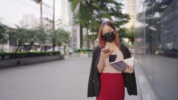 vrouwelijke kantoormedewerker draagt een medisch masker met behulp van mobiele telefoon praten met klanten, problemen oplossen via de telefoon, een secretaresse met papieren werkdocumenten, slimme zakelijke kleding, moderne stad stedelijk video