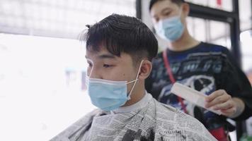 jovem asiático usa máscara facial cortando o cabelo, fica protegido e seguro fora de casa durante a pandemia covid19, barbeiro masculino cabeleireiro, higiene do cliente, sinta-se seguro. higiene da barbearia