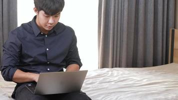 jeune homme asiatique travaillant avec un ordinateur portable assis au bord du lit, concept de travail à la maison, isolement à la maison, célébrer après avoir réussi son travail, opportunité de candidature en ligne, envoi d'e-mails video