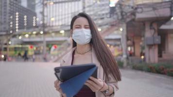 asiatische Bürodame trägt Gesichtsmaske mit digitalem Tablet, während sie außerhalb des Gebäudes geht, modernes, intelligentes Leben in der Innenstadt, bequeme Wahl für Benutzer tragbarer Gadgets, die aus der Ferne arbeiten