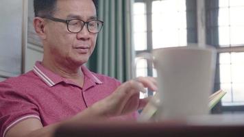 homem sênior asiático usa óculos lendo livro durante o dia dentro da sala de estar da casa, estilo de vida relaxante para pessoas de meia idade, tendo uma mente calma e pacífica, velhice aprendendo algo novo