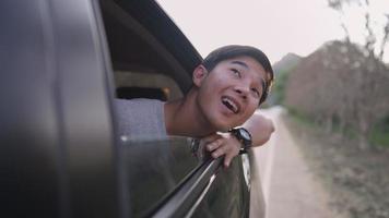 concept buiten de stad, Aziatische jonge man reist met familie in de auto, opent raam kijk naar heldere lucht, adem frisse lucht van het platteland, beweegt hand in de wind in landelijk gebied op zijweg, vakantie zomer