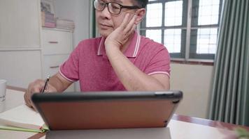 un anciano asiático usa anteojos trabajando con una tableta en el espacio de trabajo en el hogar, vejez y tecnología, presión y estrés durante el trabajo, escribiendo ideas y creatividad para personas mayores de mediana edad