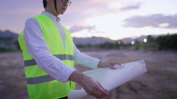 un joven ingeniero del este de Asia que usa anteojos se prepara para irse a casa, quita la mano del casco y dobla un plano de papel, trabaja horas extras dejando un sitio de construcción fangoso natural, el primer concepto de seguridad video