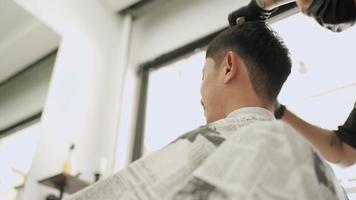 vista ad angolo basso all'interno del barbiere, il maschio asiatico si taglia i capelli. ben toelettatura, forbici per rasoio elettrico, proprietario di un barbiere durante il virus corona covid-19, riaprono le piccole imprese dopo la pandemia video