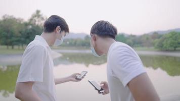 Two young men having fun talking together while relaxing in green public park, male hand showing internet information to his friend, wearing protective mask to prevent covid-19 spreading when outside video