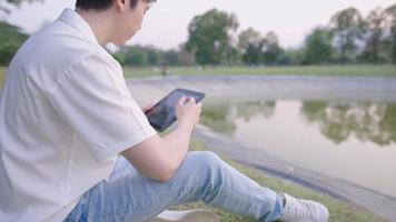 un hombre asiático solitario sentado en un amplio prado verde al lado de un estanque natural dentro del parque con las manos sosteniendo una tableta digital, buscando el concepto de inspiración, espacio de trabajo verde, aliviando la soledad video