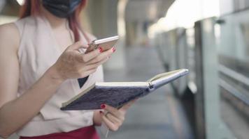 close-up shot van een jonge werkende vrouw die op het luchttreinplatform staat met behulp van een smartphone-kopie en noteerde het informatiedetail dat op het notitieboekje is geschreven, dame druk aan het werk terwijl ze op de trein wacht video