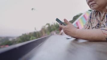 Asian modern elderly enjoy using mobile app while sitting on a long bench in natural cafe, hands sliding on social media looking at new feeds of her friends while relaxing, elderly and technology video