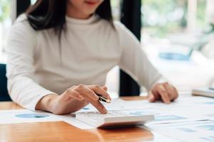 cerca de la mano del empresario o contador mano sosteniendo la pluma trabajando en la calculadora para calcular datos comerciales, documentos contables y computadora portátil en la oficina, concepto de negocio. foto