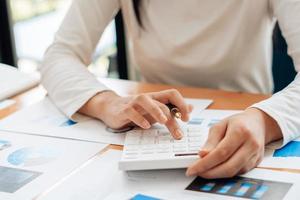 cerca de la mano del empresario o contador mano sosteniendo la pluma trabajando en la calculadora para calcular datos comerciales, documentos contables y computadora portátil en la oficina, concepto de negocio. foto