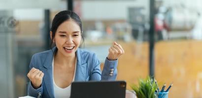 Happy asian woman office worker feeling excitement raising fists celebrates career ladder promotion or reward, businesswoman sitting at desk receive online news, great results successful work concept.