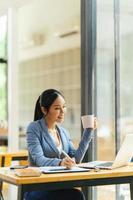 Beautiful young asian business woman drinking coffee and using laptop computer while working in office. photo