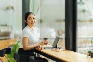 Beautiful young asian business woman drinking coffee and using laptop computer while working in office. photo