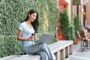 Portrait of beautiful Asian woman sitting outdoors at coffee shop restaurant during summer, using smart wireless technology computer laptop and smartphone, relaxing coffee break at cafe restaurant.