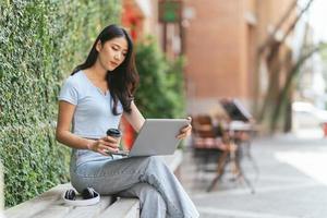 Portrait of beautiful Asian woman sitting outdoors at coffee shop restaurant during summer, using smart wireless technology computer laptop and smartphone, relaxing coffee break at cafe restaurant.
