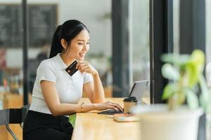 smiling woman paying online, using laptop, holding plastic credit card, sitting on coffee shop, Asian female shopping, making secure internet payment, browsing banking service. photo