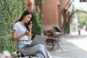 retrato de una hermosa mujer asiática sentada al aire libre durante el verano, usando una computadora portátil y un teléfono inteligente con tecnología inalámbrica inteligente, relajándose en un café restaurante. foto