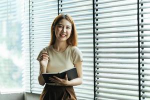 una joven mujer de negocios asiática está parada en un escritorio y tomando notas en un cuaderno. el concepto de educación y tecnología.
