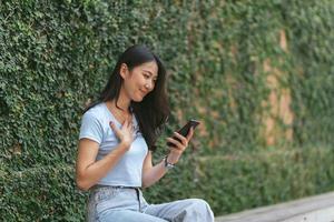 mujer asiática feliz sonriendo y sosteniendo el teléfono celular mientras se sienta en la cafetería de verano de la calle. foto