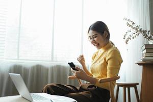 mujer joven con teléfono inteligente expresando entusiasmo en la oficina del hogar, mujer asiática emocionada se siente eufórica leyendo buenas noticias en línea. foto