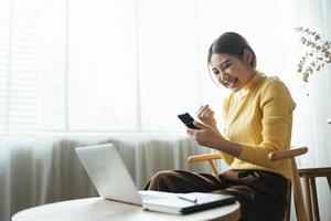 mujer joven con teléfono inteligente expresando entusiasmo en la oficina del hogar, mujer asiática emocionada se siente eufórica leyendo buenas noticias en línea. foto
