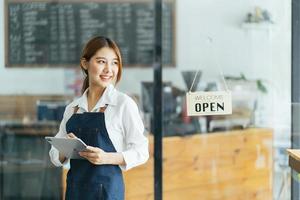 Portrait of happy woman standing at doorway of her store. Cheerful mature waitress waiting for clients at coffee shop. Successful small business owner in casual wearing blue apron standing at entrance photo